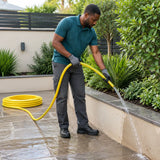 African American adult guiding a Berry yellow braided PVC hose while washing soil from a paved courtyard edge.