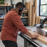African American carpenter wearing a black Climax behind-the-neck earmuff while guiding timber through a stationary table saw in a professional workshop.