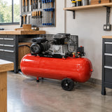 Mirage red 100L electric air compressor parked beside a workbench in an organised woodworking workshop.
