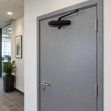 Black Mister hydraulic door closer installed above a closed office corridor door.
