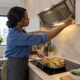 African American home cook adjusts the Kaiser stainless steel built-in hood while preparing a shallow-pan meal in a modern apartment kitchen.