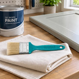 Total 1-inch oil paint brush resting on a cloth beside a closed paint tin and prepared wood panel in a home decorating station
