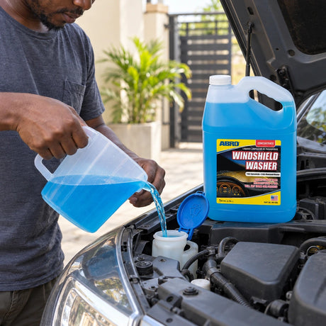 Ghanaian car owner pouring diluted blue washer fluid into a vehicle reservoir beside an Abro concentrate jug