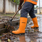 Ghanaian grounds worker wearing orange Coofix rain boots while clearing wet leaves beside a muddy drain
