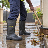 Ghanaian worker wearing black Coofix rain boots while sweeping wet leaves outdoors
